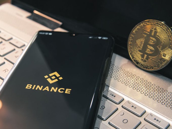 A smartphone displays the Binance logo on its screen, resting on a laptop keyboard next to a physical Bitcoin token.