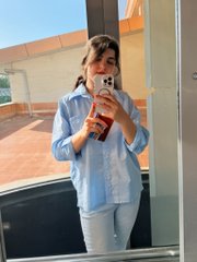 A person takes a mirror selfie wearing a light blue button-up shirt and white pants, holding a phone with a white polka-dot case.