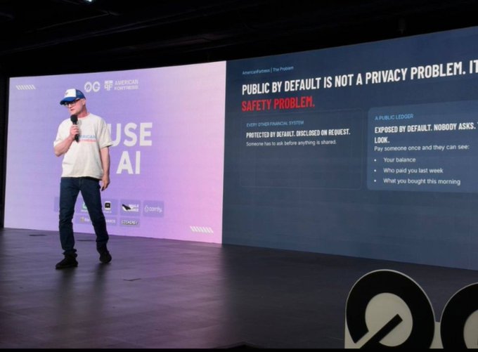 A speaker on stage wearing a white t-shirt, cap, and sunglasses presents a slide titled 'PUBLIC BY DEFAULT IS NOT A PRIVACY PROBLEM. IT'S A SAFETY PROBLEM.' at an AI conference.