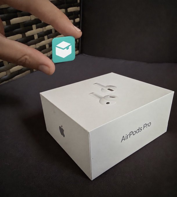 A hand holds a small teal cube with a white graduation cap icon above an AirPods Pro box resting on a dark surface.