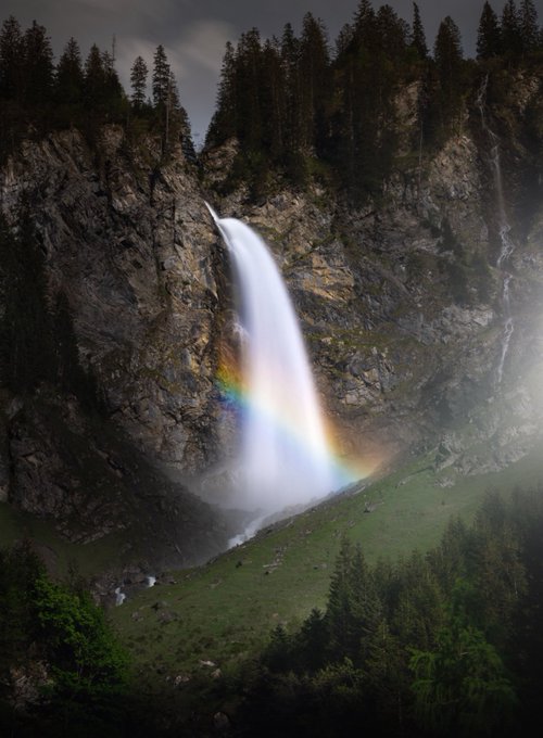 A tall waterfall cascades down a rocky cliff into a green valley, forming a vivid rainbow in its mist at twilight.
