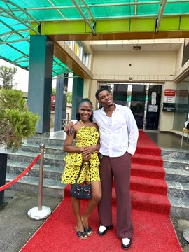 A well-dressed couple poses happily together on a red carpet in front of a building entrance, smiling for the camera with the woman in a yellow floral dress and the man in a white shirt and brown trousers—capturing the joyful confidence the post references about overcoming the fear of rejection.