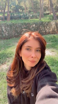 A selfie photo of a young woman with long brown hair wearing a dark hoodie, taken outdoors in a green, tree-lined area.