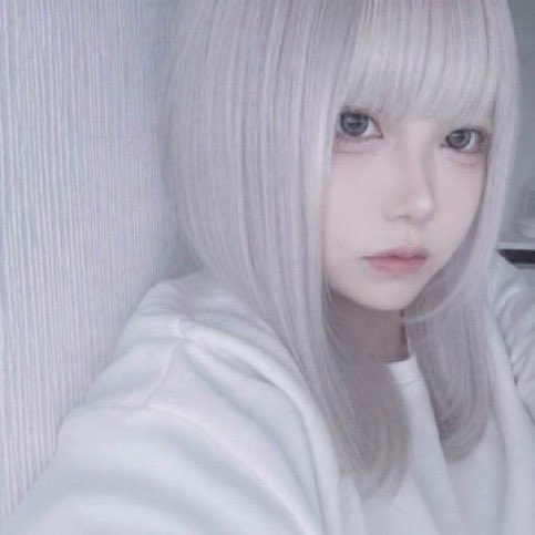A young woman with long silver-white hair and bangs poses in a selfie-style portrait, wearing a white top against a pale background.