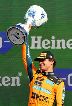 A McLaren Formula 1 driver in an orange papaya race suit triumphantly lifts the Epsom-sponsored trophy above his head on the podium, with "OKX" branding visible on his chest, celebrating their championship victory.