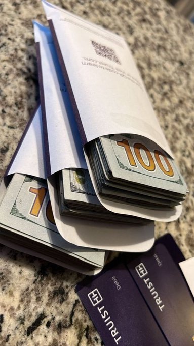 Two white envelopes filled with stacks of $100 bills sit on a granite countertop next to two purple Trust Wallet cryptocurrency cards.