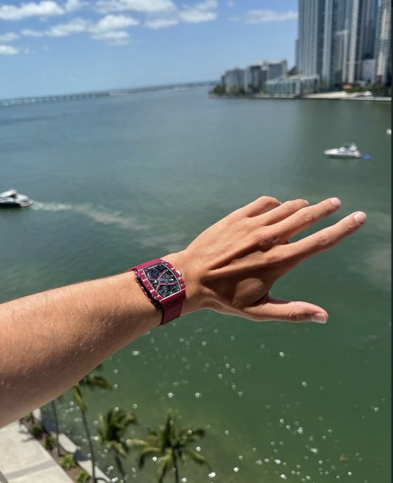 The image features a person wearing a red wristwatch, with their hand extended outwards towards the camera. In the background, there are several boats floating on the water near some buildings. The photo also includes text that reads "1.5 $SOL Just hit the like and say gm".