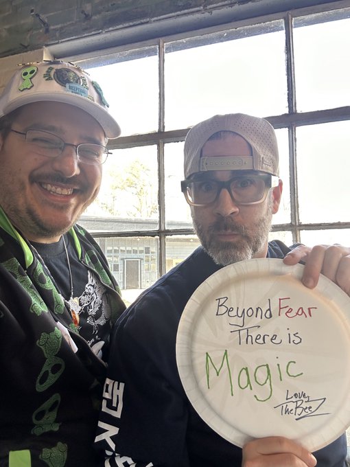 Two men posing for a photo; the man on the right is holding a frisbee with the handwritten message 'Beyond Fear There is Magic - Love, The Bros'.