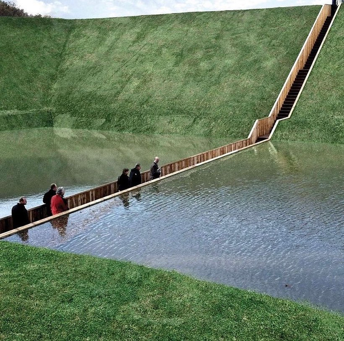 A wooden walkway partially submerged in water, with people standing on it within a grassy, excavated landscape.