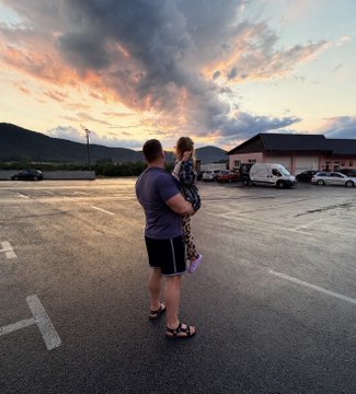 Two people standing in a parking lot at sunset, watching a colorful sky with mountains and buildings in the distance.