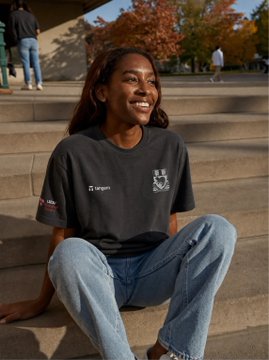 A young woman smiles while sitting on campus steps wearing a black Tangem x LSE t-shirt, capturing the launch of Tangem Campus at the London School of Economics to promote self-custody education among student builders.