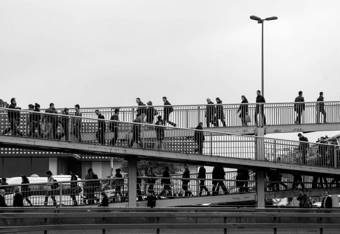 A black-and-white photograph captures pedestrians walking across a multi-level urban pedestrian bridge with metal railings, street lamps, and onlookers on various tiers.