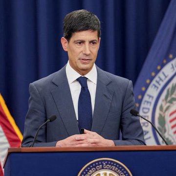 A man in a dark suit and blue tie stands at a podium bearing the seal of the Federal Reserve Board of Governors, flanked by U.S. and Federal Reserve flags, suggesting an official Federal Reserve address. The setting aligns with the post's discussion of upcoming PCE inflation data and potential implications for Fed monetary policy decisions.