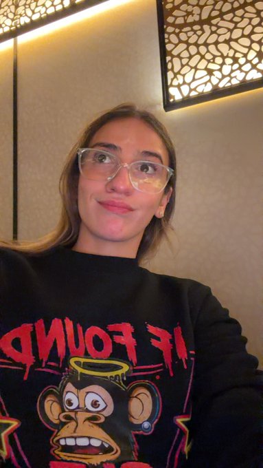 A young woman with clear-framed glasses and a black sweater featuring a cartoon monkey with a halo poses for a selfie in a room with decorative lampshades.