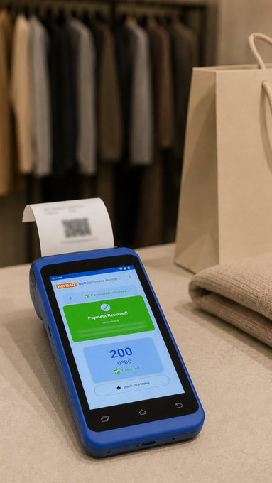 A blue payment terminal displays 'Payment Received' and '200 USDC' next to a printed receipt, with a clothing rack and shopping bag in the background.