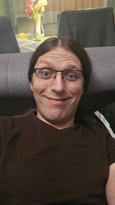 A woman with glasses and dark hair offers a reluctant but amused smile from a couch, embodying the "feeling lazy but knowing it's time to grind" mood of the Wednesday morning post. A small white name tag with "Roxanne" is visible on her shirt.