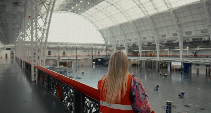 A woman with long blonde hair, wearing an orange safety vest over a patterned top, looks out over a large, mostly empty concourse, likely a train station.