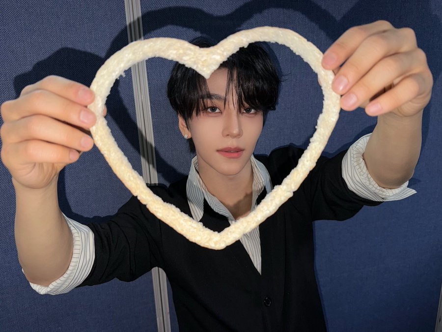 A young man with dark hair holds up a large heart-shaped rice cake in front of his face, dressed in a striped shirt and black cardigan against a blue backdrop. The playful, affectionate gesture complements the fan-engagement tone of the "#ALPHADRIVEONE" and "#XINLONG" hashtag update, suggesting a promotional or celebratory post for the artist or group.