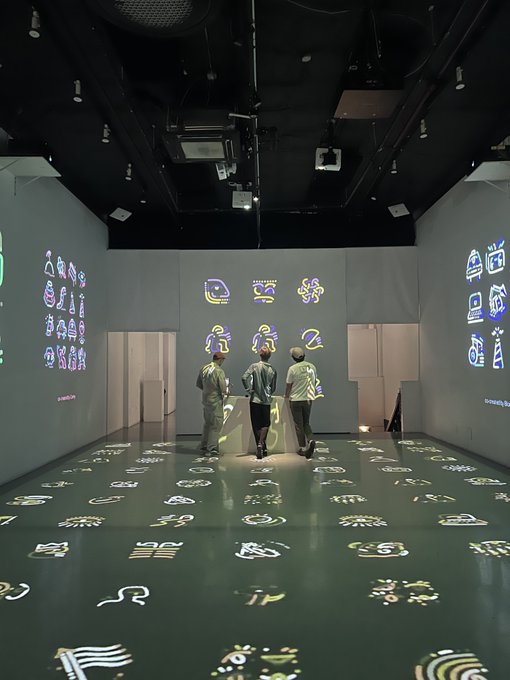 Three people stand in a dimly lit exhibition room with abstract symbol projections covering the walls and floor, exploring an immersive art installation.