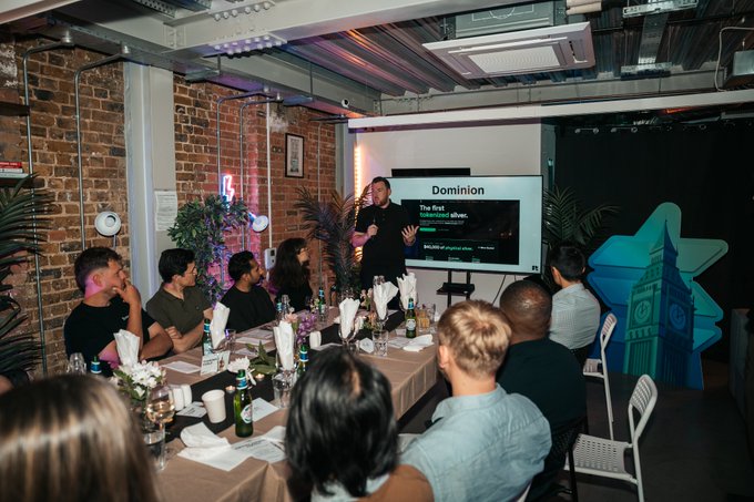 A diverse group of people seated around a long dining table listen to a presenter giving a talk titled 'Dominion: The First' at what appears to be an intimate tech meetup or workshop.