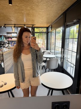 A young woman with long brown hair takes a mirror selfie in what appears to be a café, wearing a white lace top and shorts under an oversized denim jacket, smiling while holding her phone. The casual, stylish photo aligns with the post's travel theme, capturing a relaxed pre-trip moment before her 21-hour journey.