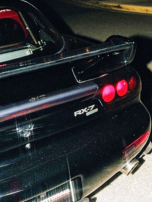 A black Mazda RX-7 sports car is photographed from the rear at night, showcasing its large rear spoiler, illuminated red taillights, and the "RX-7" badge on the trunk. The image has a grainy, film-like quality with subtle star-like sparkles across the dark paintwork.