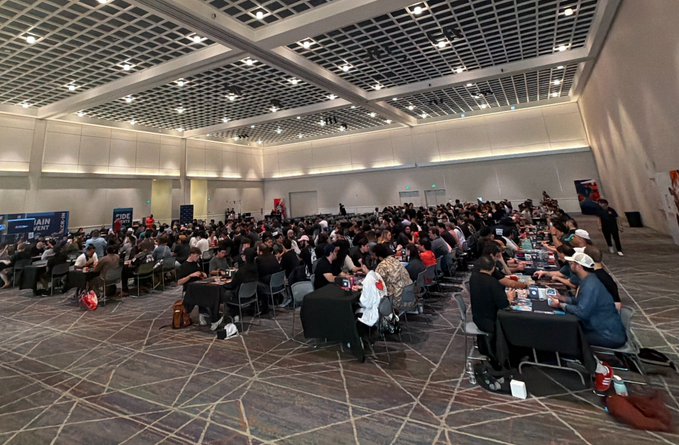 A large indoor convention hall filled with people seated at long tables, appearing to participate in a card tournament or similar event.