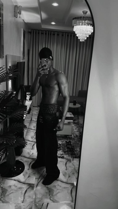 A black and white mirror selfie of a shirtless, muscular man wearing a beanie and patterned boxer briefs, standing in an elegant room with a chandelier.