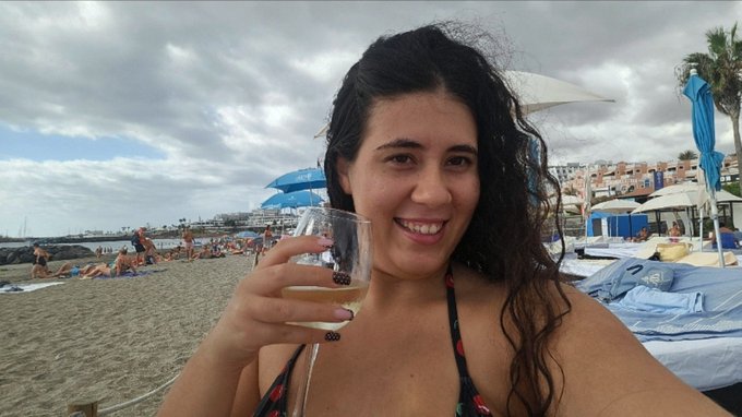 A woman with curly dark hair smiles brightly while holding a glass of white wine at a beach club, with blue umbrellas, lounge chairs, and the sea visible behind her—capturing the carefree "living my best life" Saturday vibe described in the post.