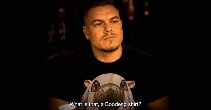 A man wearing a black Moodang t-shirt featuring a frog graphic appears in a dimly lit interview setting, with the subtitle "What is that, a Boodang shirt?" questioning the design and prompting his amused reaction.