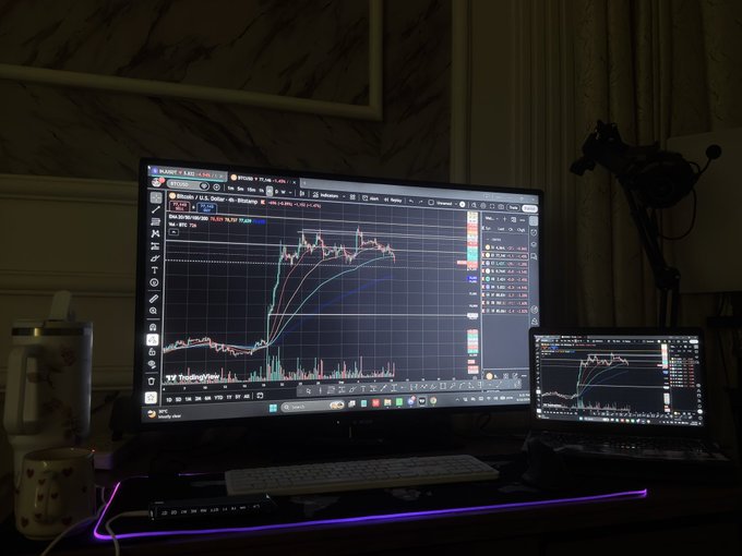 A dimly lit desk setup showing a large monitor and laptop displaying the TradingView platform with active cryptocurrency price charts (showing Bitcoin/FDUSD) and volume indicators.