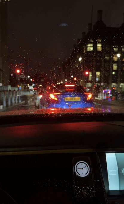 A nighttime view through a rain-speckled windshield of a car stopped at a red light behind a blue car with illuminated brake lights, showing a dashboard clock at 9:00.