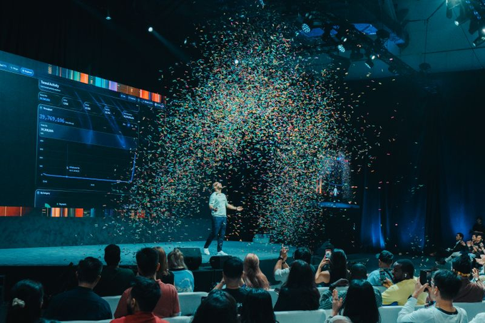 A person standing on stage at a conference or event with colorful confetti exploding around them, while an audience takes photos and a large screen displays data in the background.