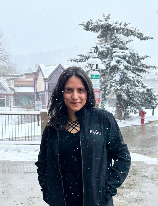 A woman in a black jacket with 'PYNS' branding standing outdoors in a snowy setting with a snow-covered tree and street lamp in the background.