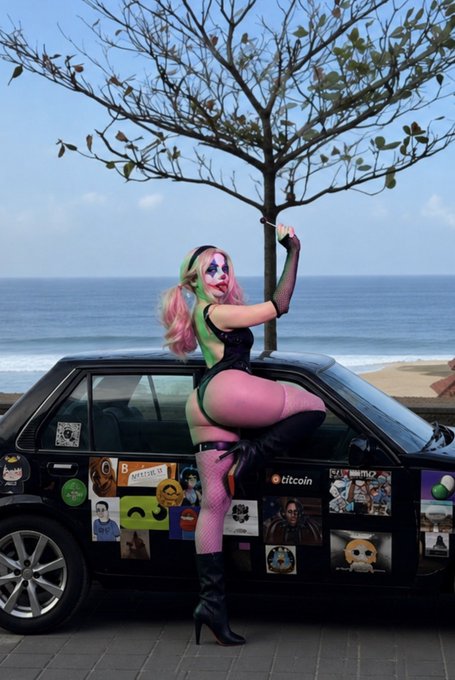 A woman with clown makeup, dressed in a gothic, playful outfit, strikes a pose on a car covered in stickers, set against an oceanic backdrop, rebelling against traditional standards of beauty. This image aligns with the person in the caption who goes by the name of $Clussy GCR and is tagging the person who goes by the name @1somniac2death who could be an artist, curator, or influential person within digital or creative spheres as they are being tagged it is highly probable a reply to them as a reply the response may relate to creative output, collaboration, or recognition.