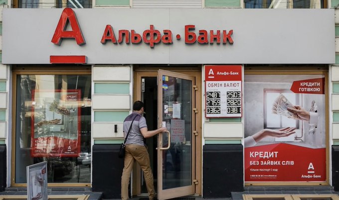 A man entering an 'Альфа-Банк' (Alfa-Bank) branch with currency exchange rates displayed on the window and an advertisement reading 'КРЕДИТИ ГОТІВКОЮ' and 'КРЕДИТ БЕЗ ЗАЙВИХ СЛІВ'.