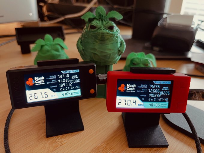 Two handheld cryptocurrency mining devices (HashCash Consultants) display real-time mining statistics, with green figurines visible in the background.