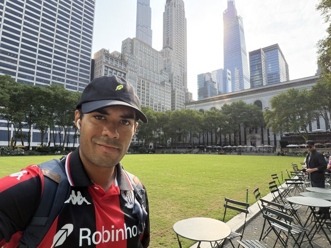 A selfie of a man wearing a black Robinhood cap and a red and black Kappa soccer jersey with 'Robinhood' sponsor text, taken in a city park with skyscrapers in the background.