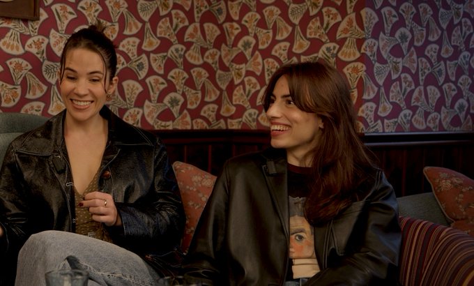 Two smiling young women sitting on a couch in front of butterfly-patterned wallpaper; one wears a black leather jacket over a gray top, and the other wears a black leather jacket over a printed t-shirt.