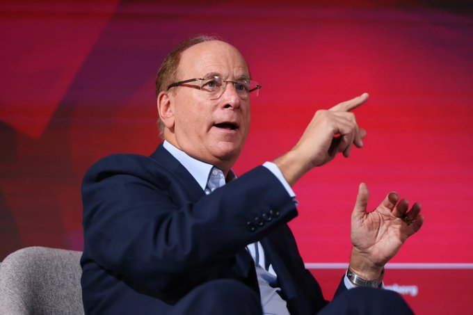 An older man in a dark suit and glasses gestures with his hand while speaking on a stage with a red background.