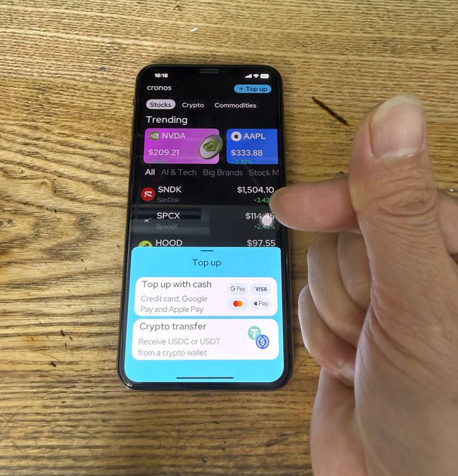 A hand is shown interacting with a smartphone displaying the "cronos" app, which features trending stocks like NVDA ($209.21) and AAPL ($333.88), with a pop-up menu offering options to "Top up with cash" via credit card, Google Pay, and Apple Pay, or use a "Crypto transfer."