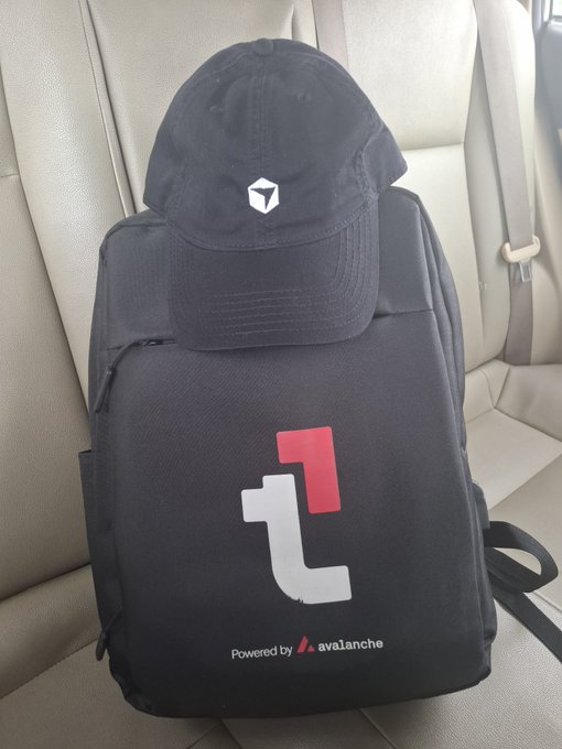 A black backpack and matching cap featuring the Avalanche (AVAX) logo are displayed on a car seat, with the backpack labeled "Powered by Avalanche" and the cap branded with the Cursor AI logo—showcasing collaborative merchandise from the Cursor AI x Avalanche Team1 partnership.