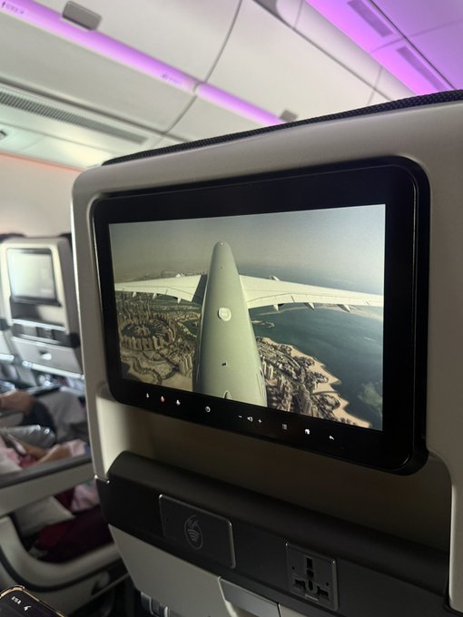 A passenger on a flight relaxes by watching a live tail-mounted camera feed on the seatback entertainment screen, showing the aircraft's wing and tail fin soaring above a coastal landscape. The scenic aerial view perfectly illustrates the post's "Dead Price Action = Vacation Time" sentiment, as the trader takes a break from flat markets to enjoy a well-earned getaway.