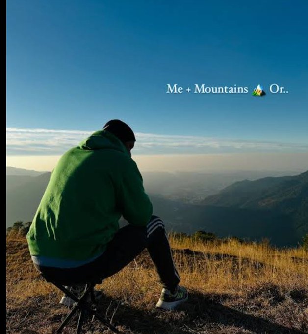 A person in a green hoodie sits on a stool on a mountain overlooking a vast landscape, with the text 'Me + Mountains 🏔️ Or..' overlaid on the sky.