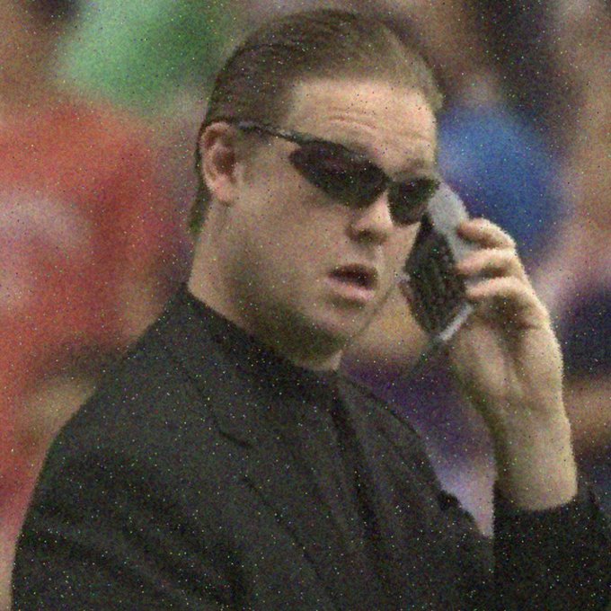A grainy photo of a muscular man wearing dark sunglasses and a black shirt, talking on a silver flip phone with a blurred crowd in the background.