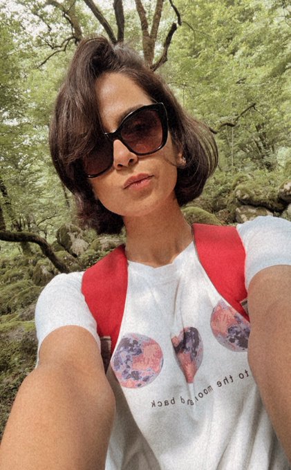 A selfie of a woman with sunglasses and a graphic t-shirt wearing a red backpack in a lush green forest.