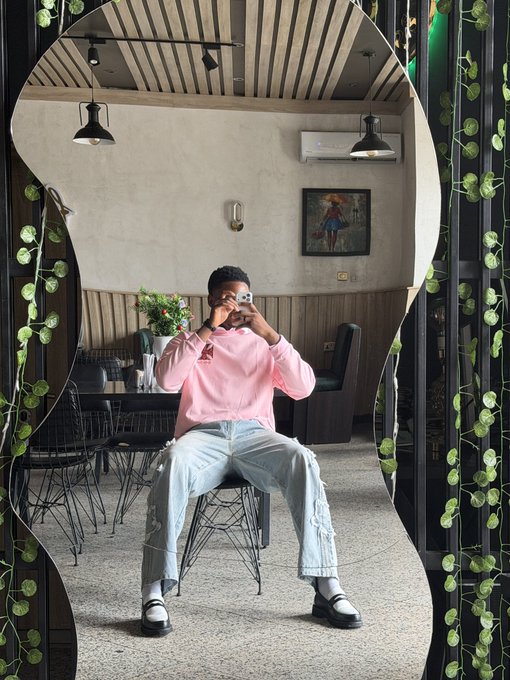 A young man wearing a pink 'A' logo sweatshirt sits on a stool taking a mirror selfie inside a stylish cafe with hanging plants and an arched mirror frame.