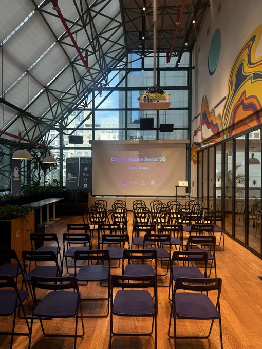 An empty event space set up with rows of black chairs facing a projection screen displaying 'Crypto House Seoul '25' and 'Warm Korea'. The venue features a high industrial ceiling with exposed beams and colorful wall art.