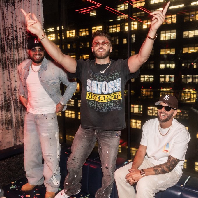 Three men posing at a nighttime party, with the center figure wearing a 'Satoshi Nakamoto' t-shirt and raising his arms.