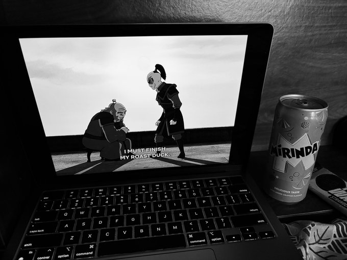 A black and white photo of a MacBook displaying an animated scene with subtitles reading "I MUST FINISH MY ROAST DUCK," next to a can of Mirinda soda.